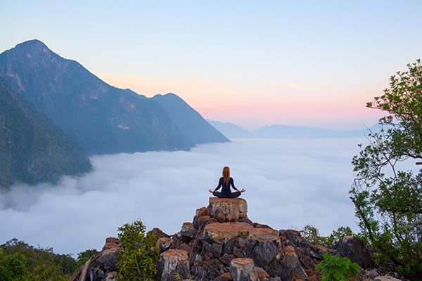 Meditierende auf einem Berg, im Hintergrund Berggipfel und Wolken über dem Tal